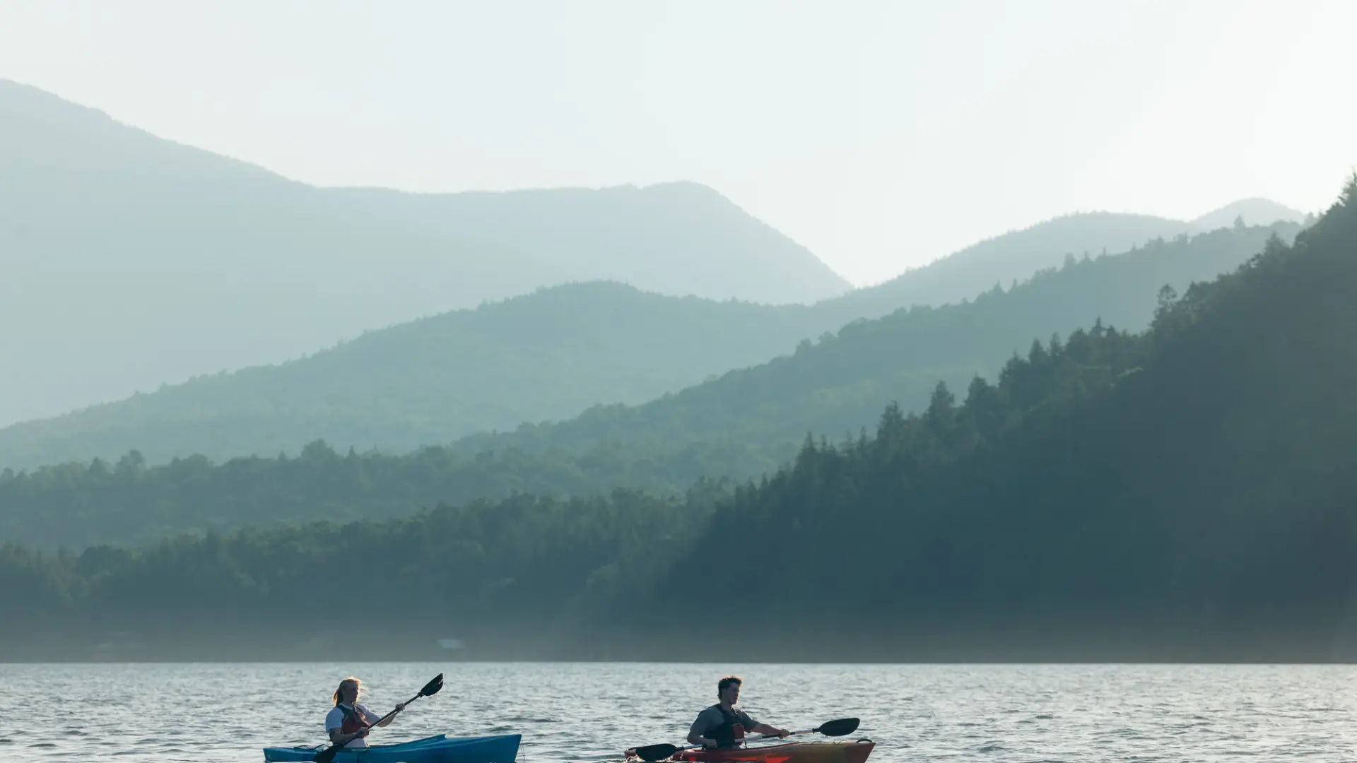 Two kayakers paddle on Lake Placid with layered mountains in misty background