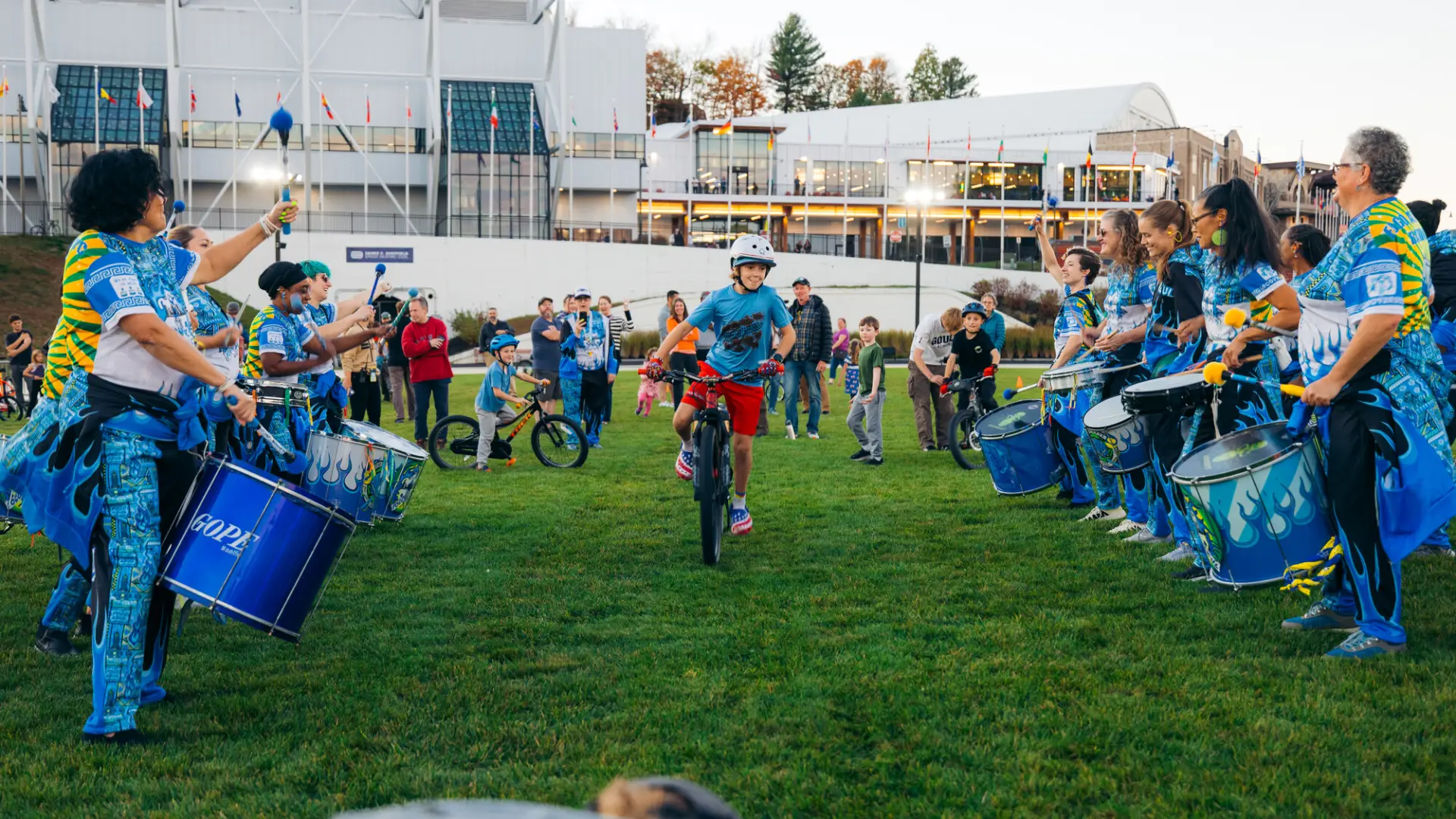 Boy rides bike between rows of a performing drum line as other kids wait their turn and adults look on at Olympic Oval