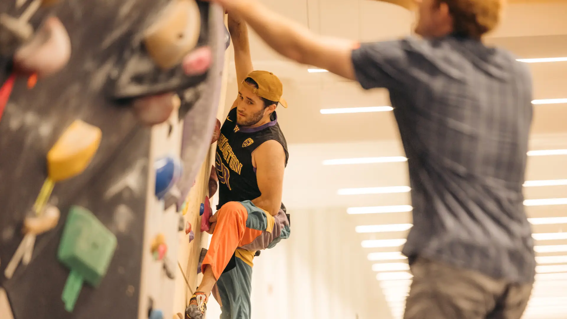 Boulderer seen climbing on indoor rock wall while another person reaching out to touch hold on wall in foreground