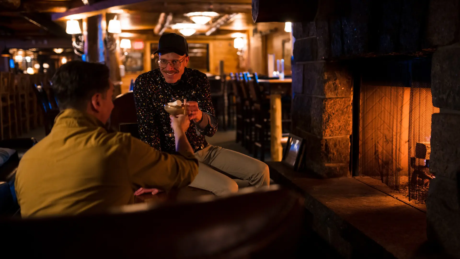 Two men enjoy drinks by a fireplace in a dark bar