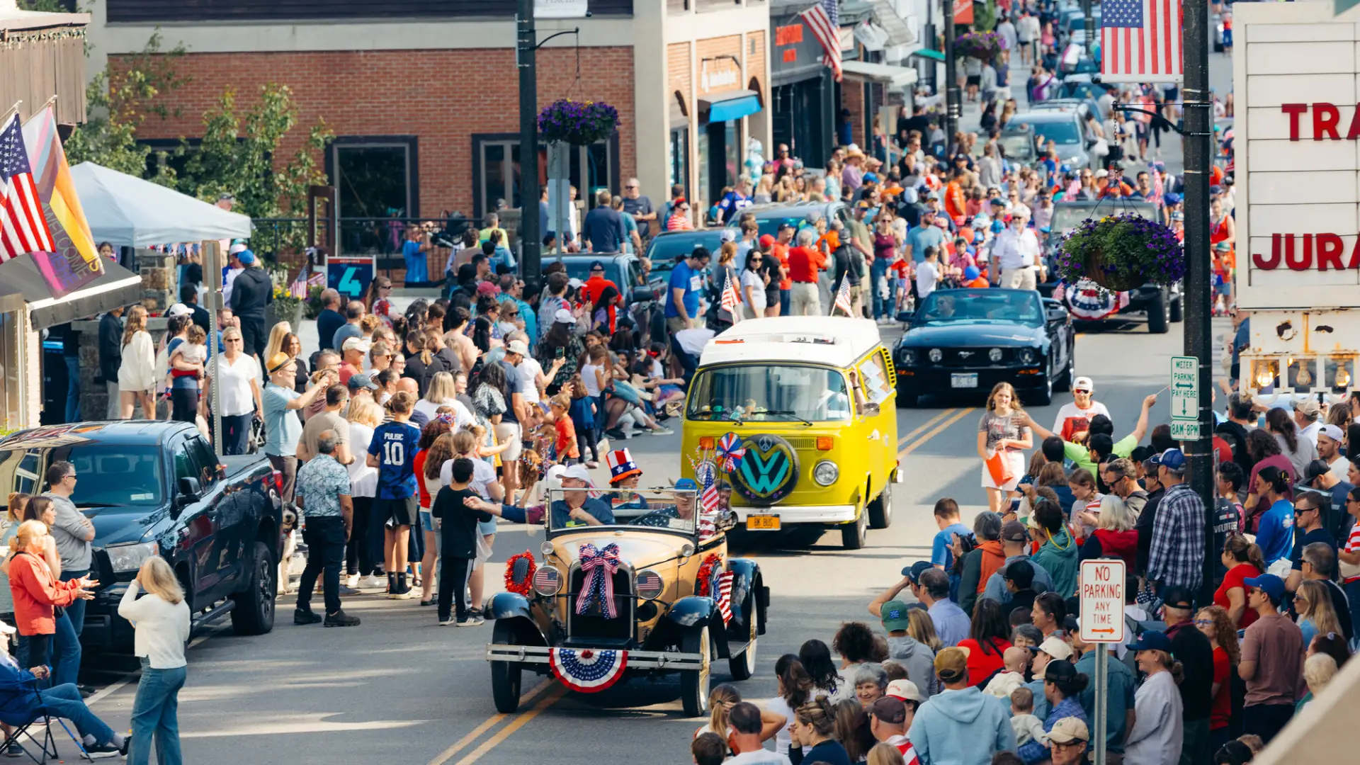 A festive Independence Day parade proceeds down a flag-lined Main Street in Lake Placid, featuring vintage cars.