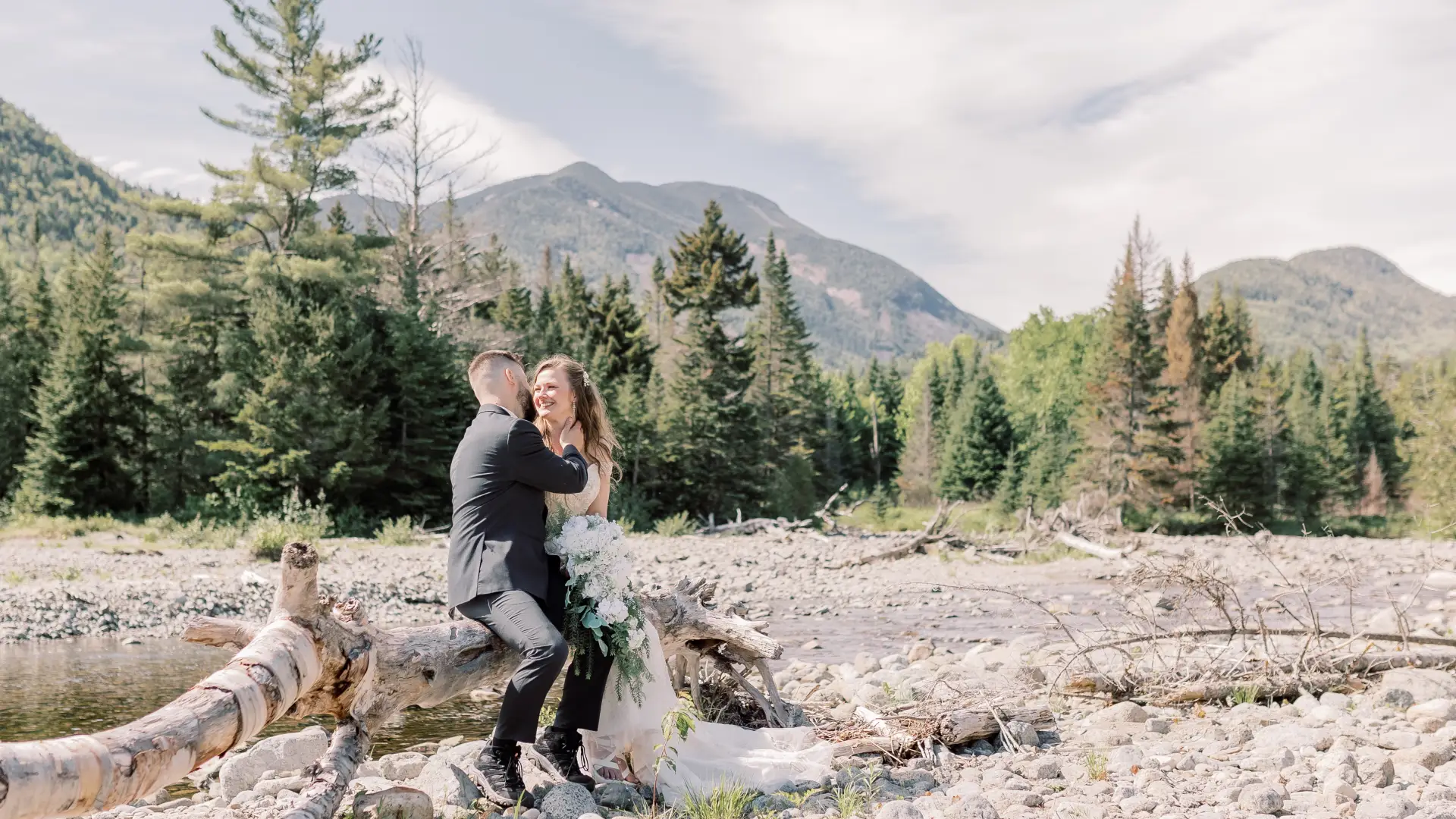 bride and groom snuggle on a fallen tree by a rocky river bed with mountain backdrop