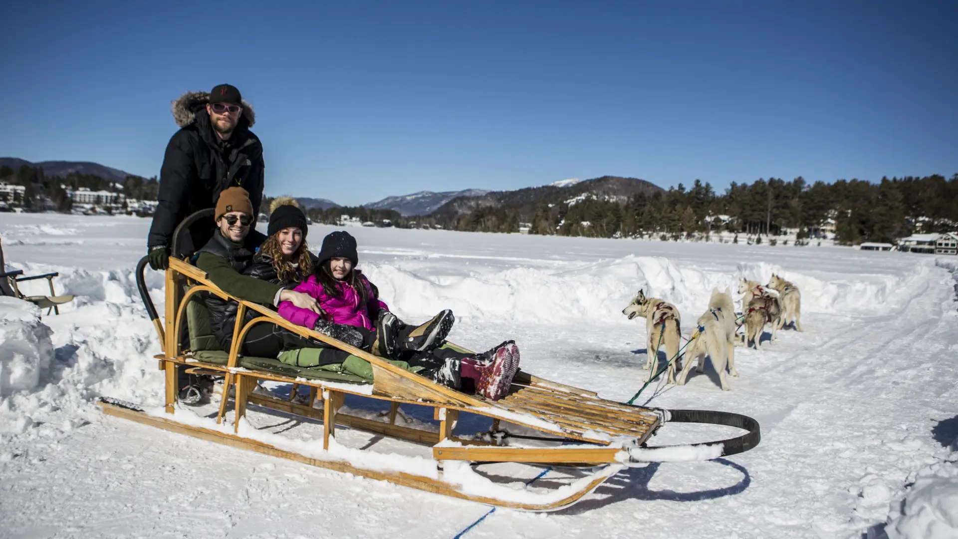 A family poses on a dog sled, with a driver standing on the back of the sled, on a frozen lake.