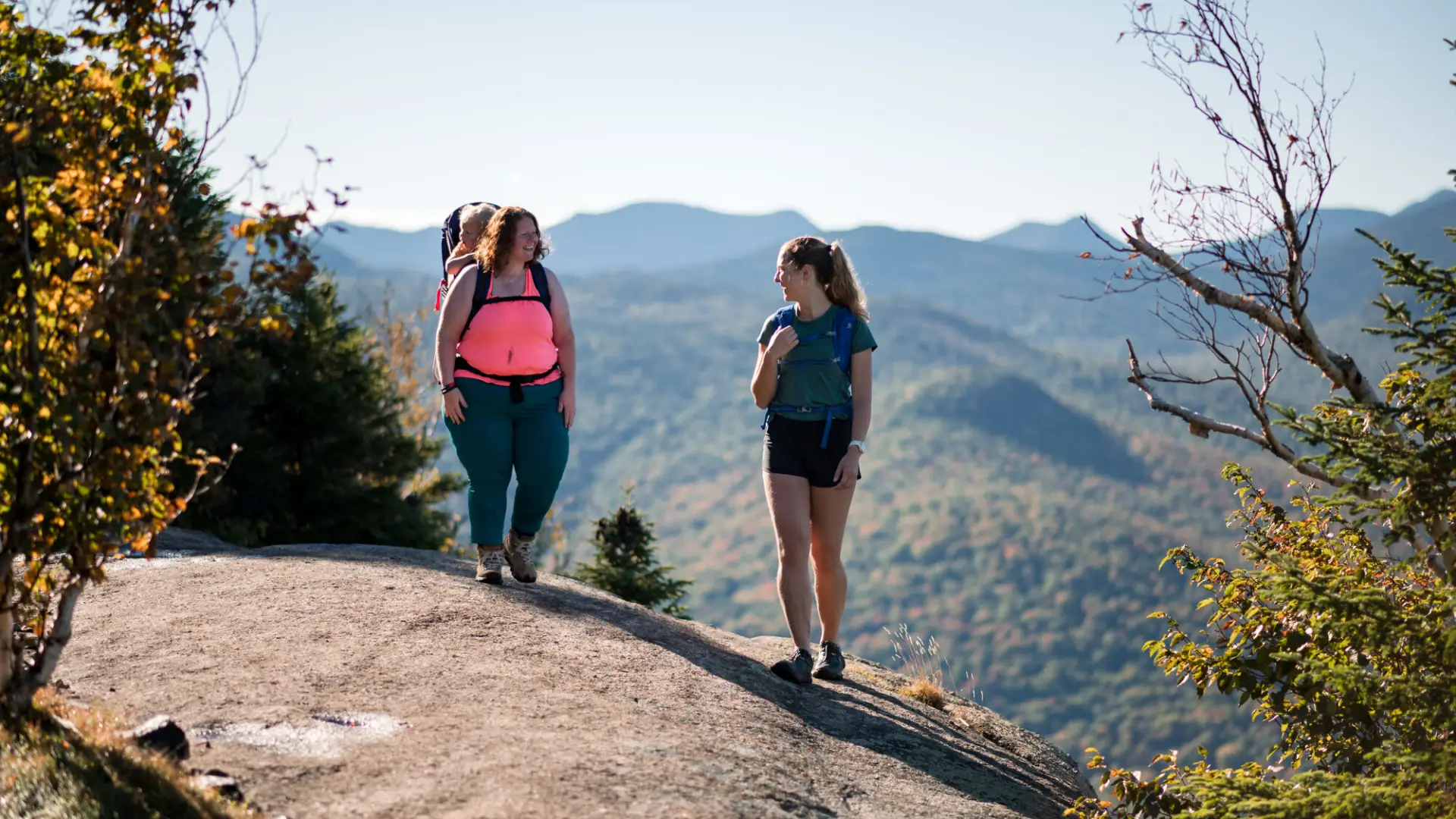 Two woman atop a mountain, one with a baby in her backpack