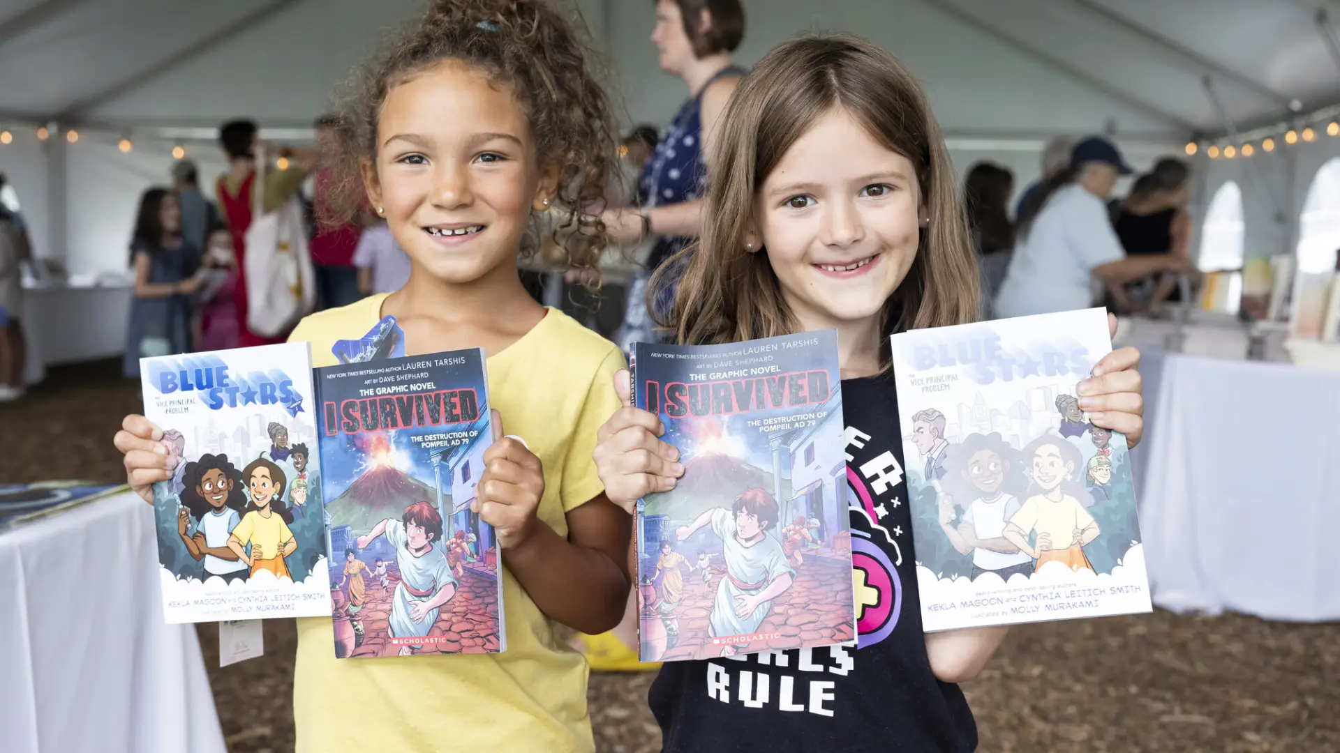 Two young girls stand side by side holding up two children's books each while smiling at the camera