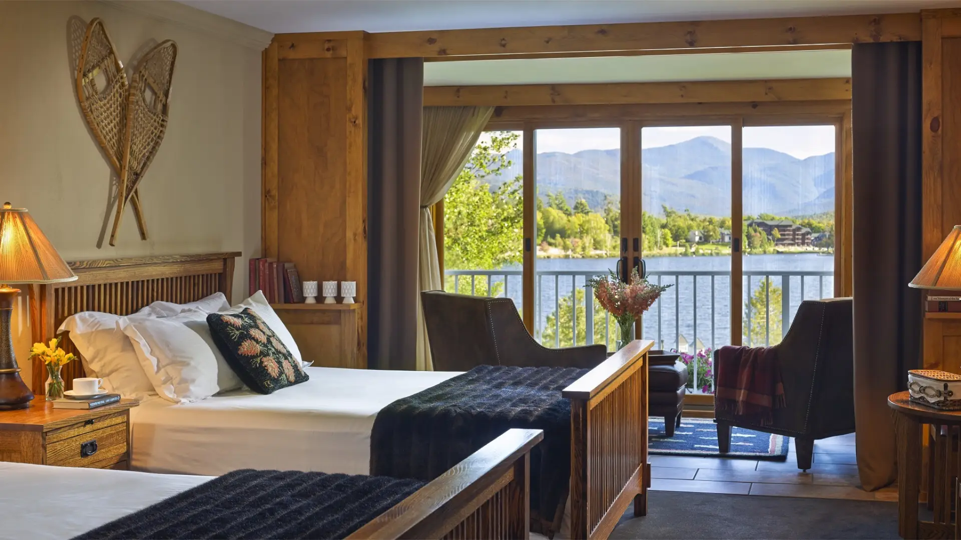 Beautifully appointed bedroom space with adirondack decor like snowshoes adorn the walls and the balcony windows open showing Mirror Lake and the majestic mountainscape background