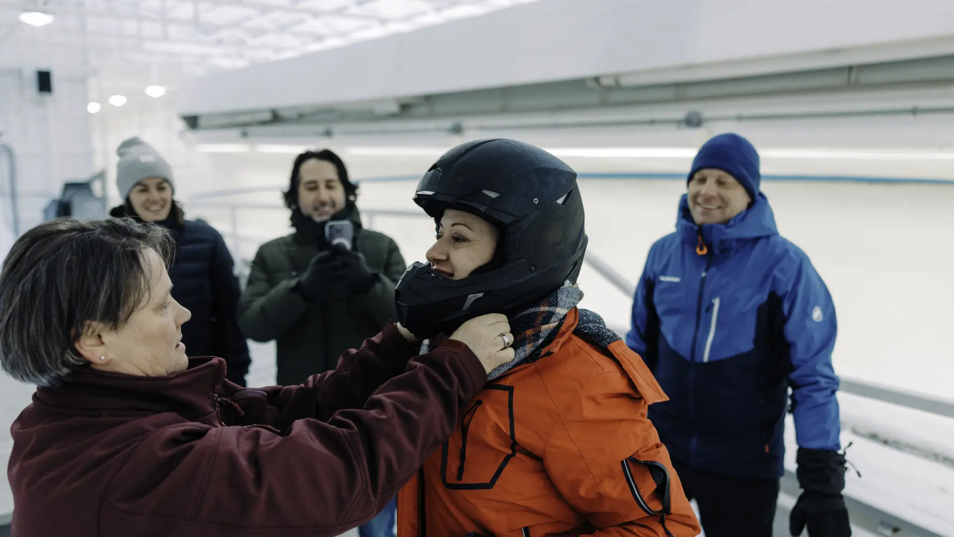Track worker helps woman put on helmet outdoors at an ice sliding track while others look on