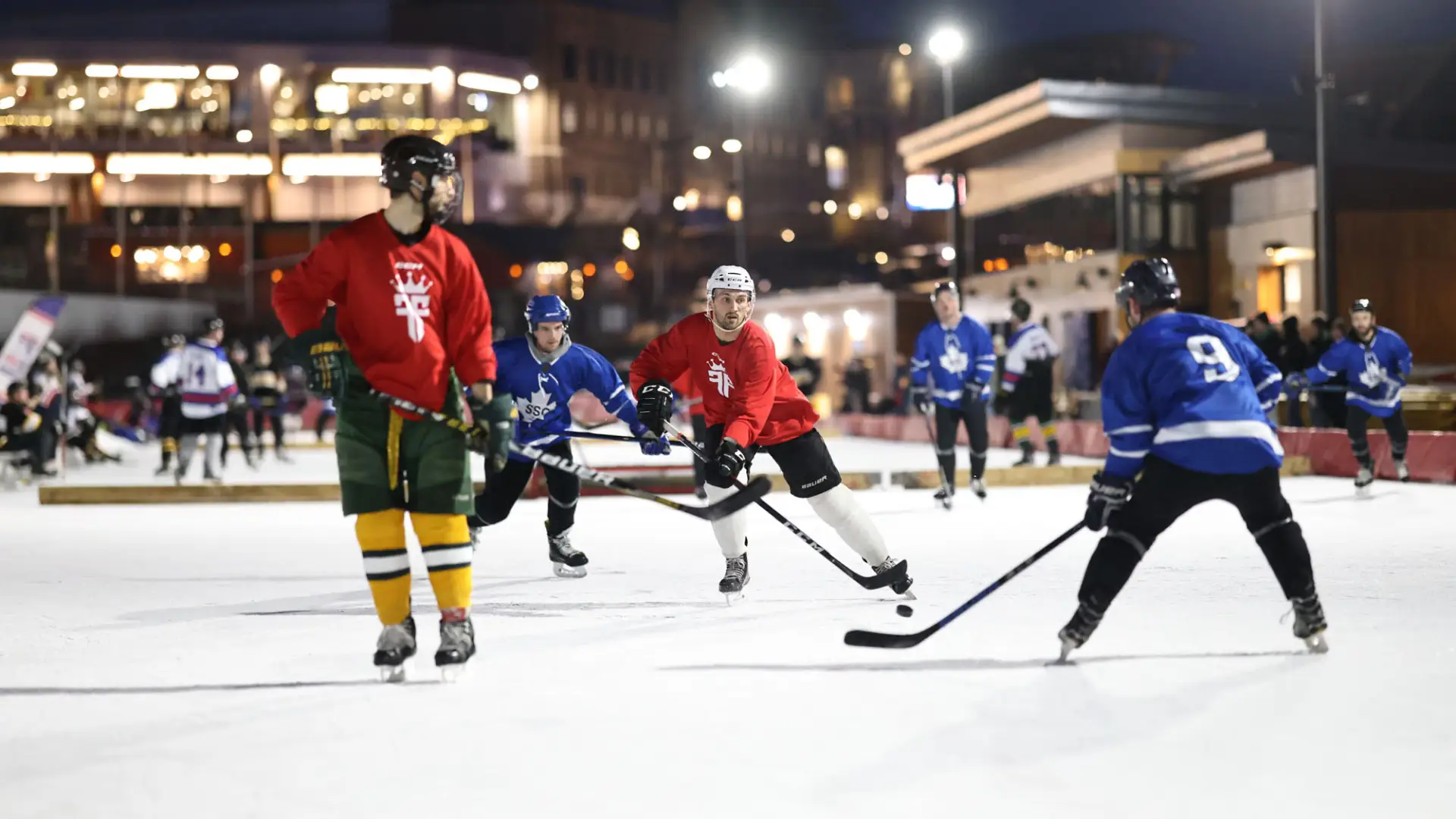 Hockey players in red and blue jerseys compete on the ice of the Olympic Oval under the lights at night