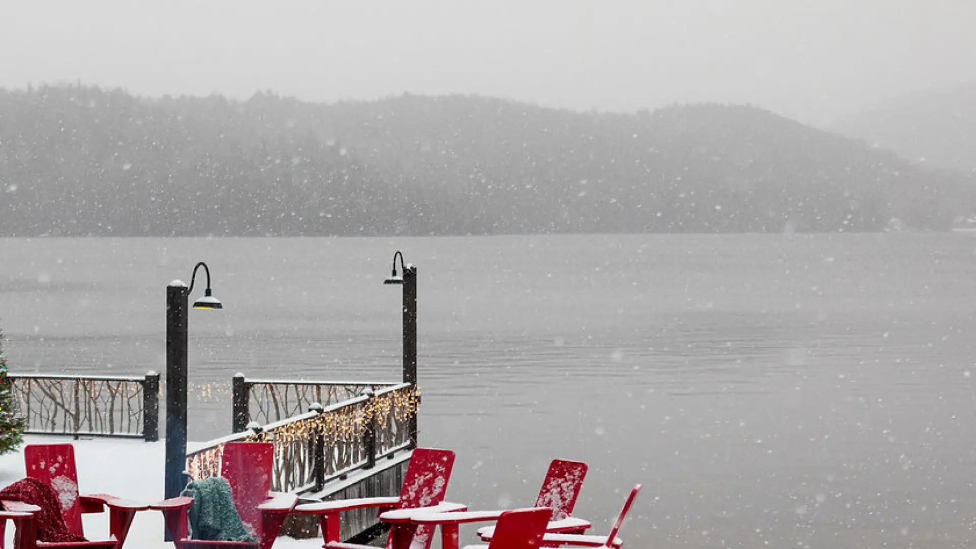 A firepit surrounded by red adirondack style chairs under light snow next to a lake.