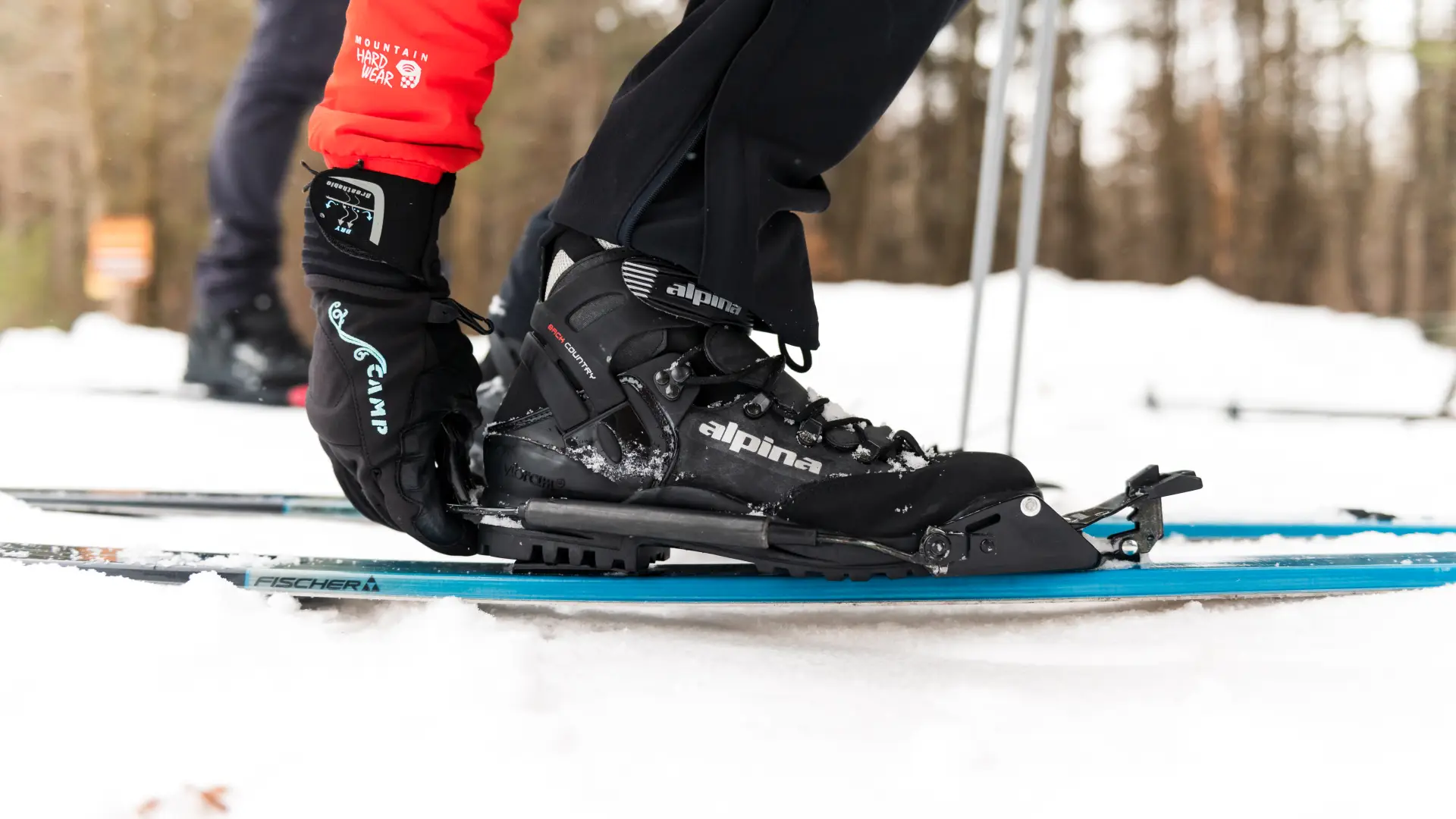 A person in red and black ski gear adjusts their boot binding on a blue ski against a snowy backdrop, conveying a sense of readiness and focus.