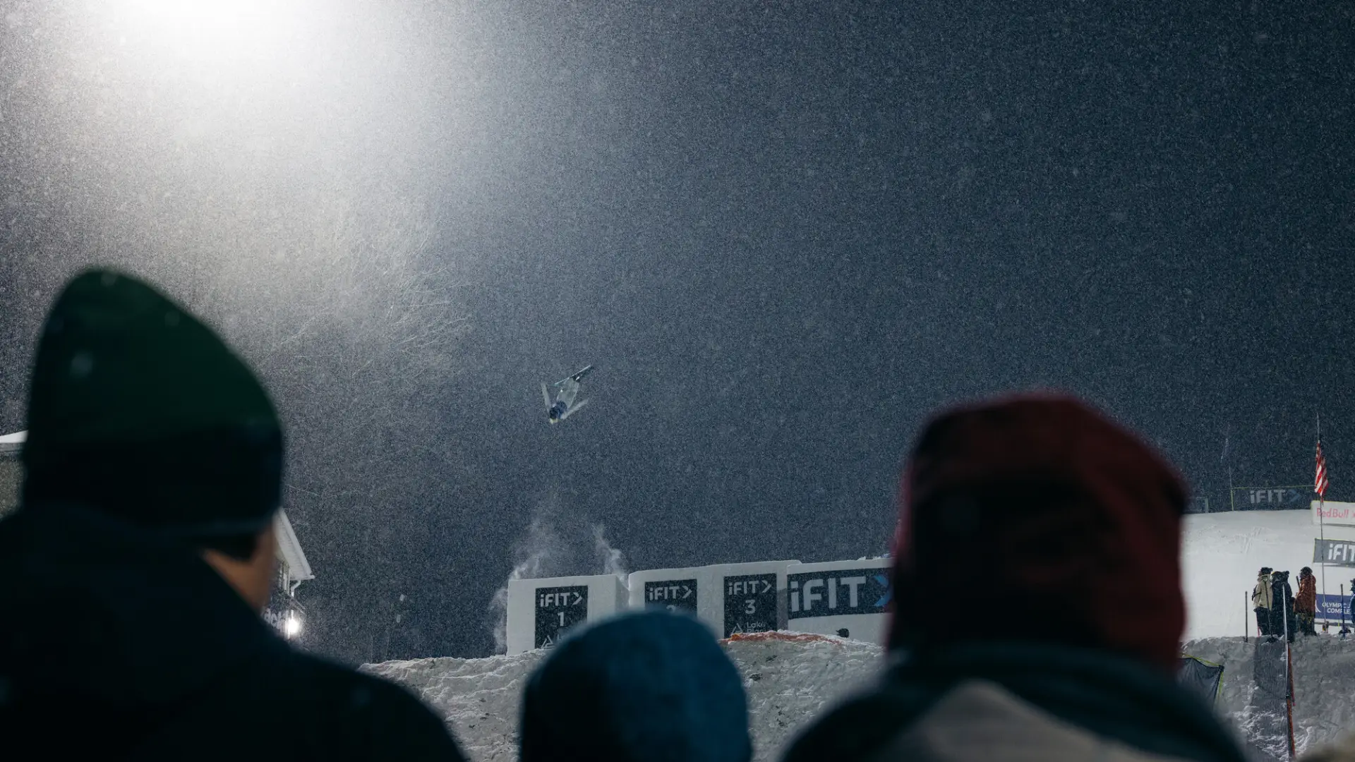 Freestyle aerial skier launches into a flip off a jump on a snowy hill at night, illuminated by artificial light. Dark heads look on in the foreground