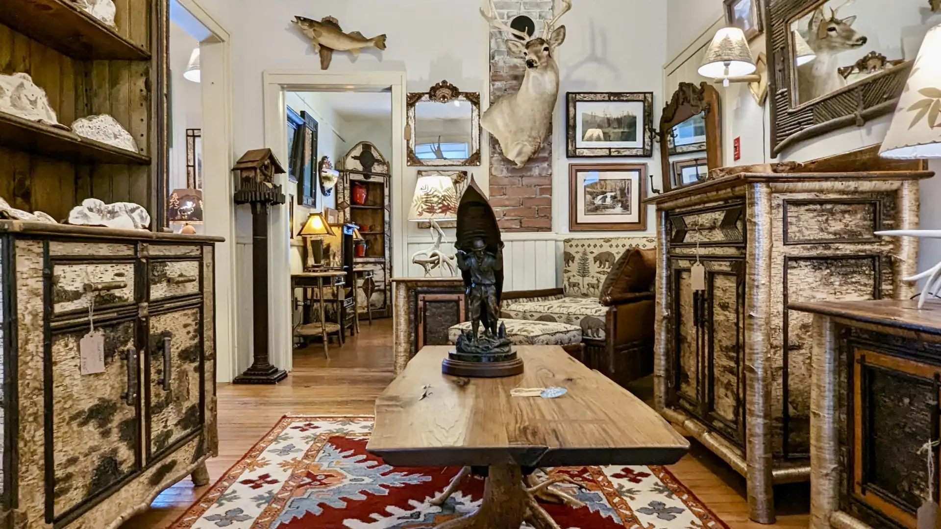 Large hutch and matching dresser made from white birch bark, a coffee table with live edge top and root ball base, a carving of a man carrying a canoe, mounts of various animals hang on the walls punctuated with mirrors surrounded with various barks.