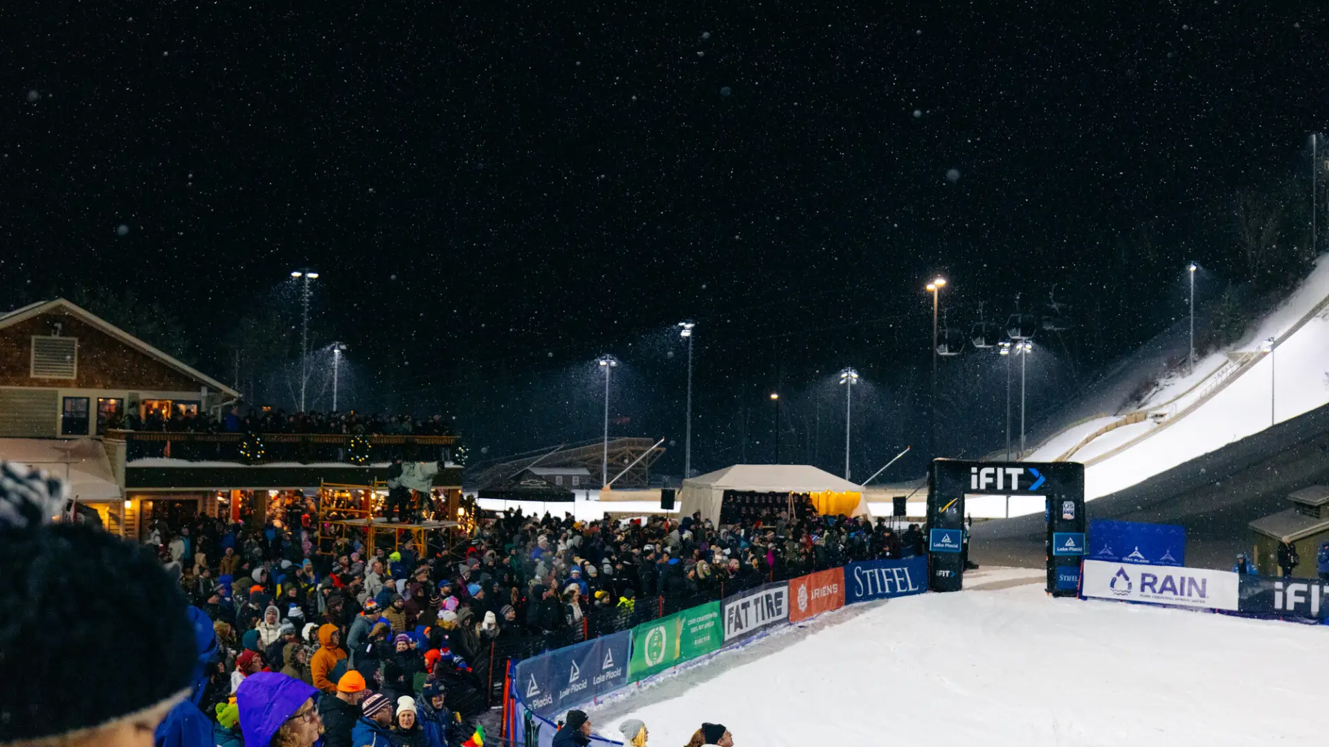 Crowd is seen in front of the Intervale Lodge at the Olympic Ski Jumps for a nighttime freestyle aerials competition. The crowd gathers around the skiers finish area, held back by a barricade.