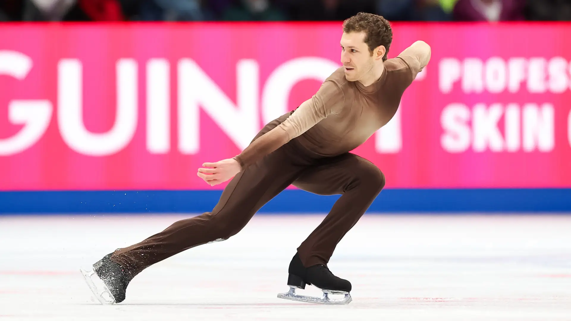 Male figure skater in brown long sleeve and long pant spandex suit performs a crouched move on ice