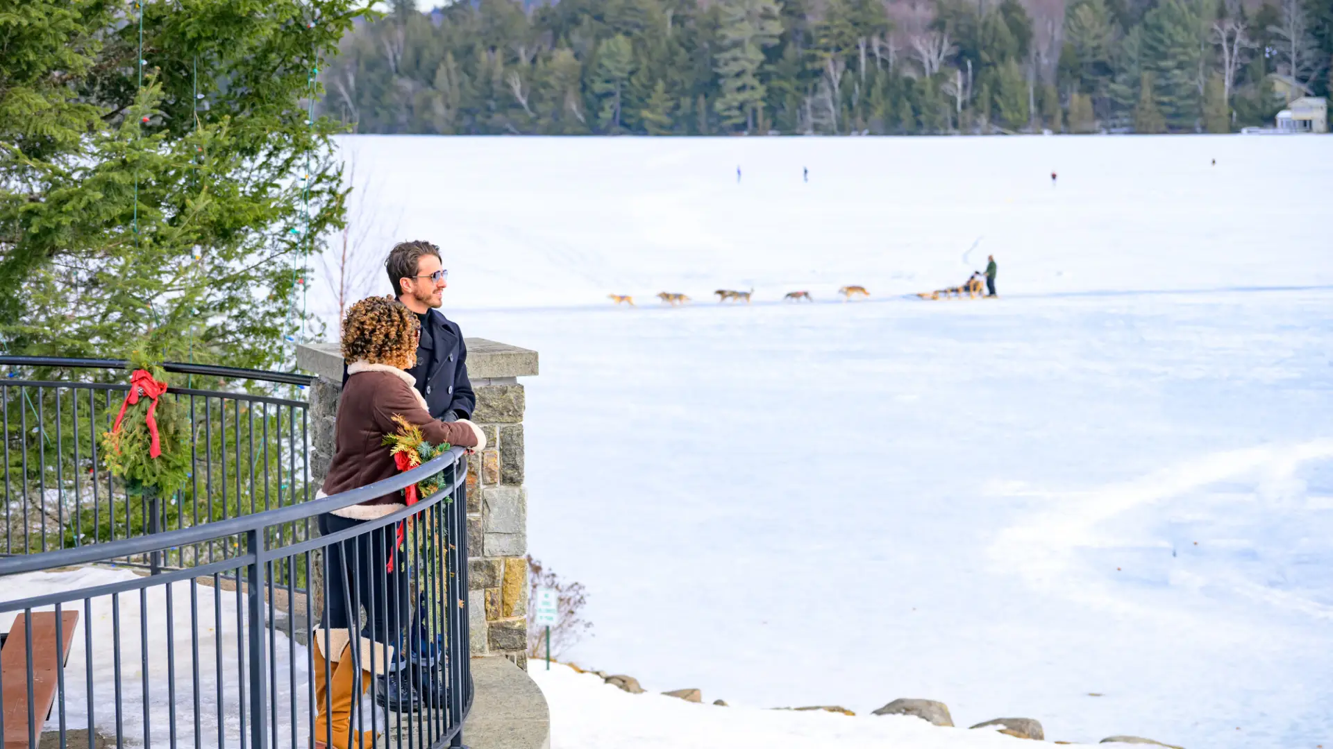 Man and woman stand on patio overlooking frozen lake with dog sleds and distant mountains