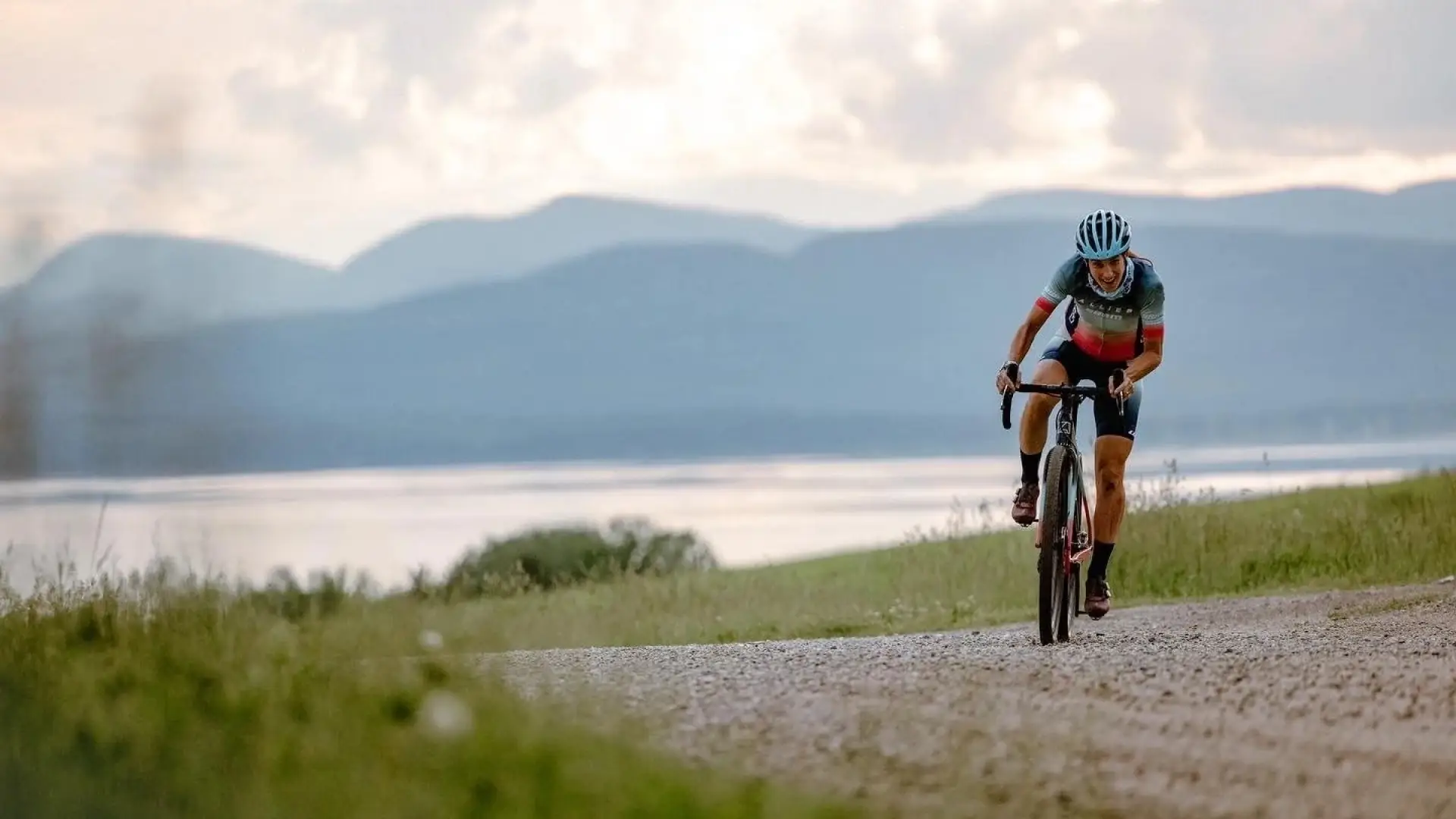 Lea Davison rides mountain bike on gravel road next to large body of water with mountains in the distance