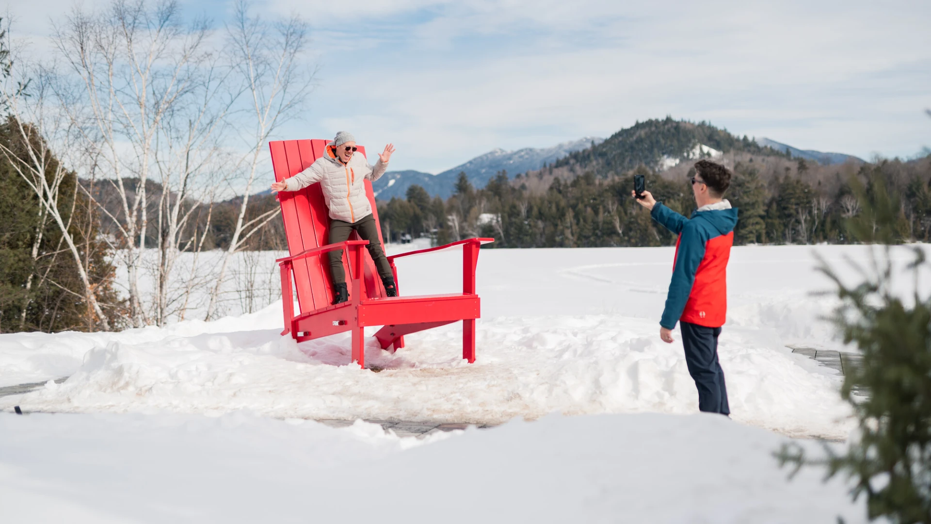 A man takes a photo of another man posing on a giant Adirondack chair in a snowy park with mountains in the background.
