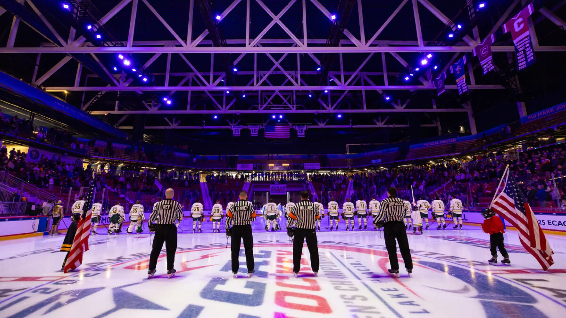 Indoor ice hockey arena filled with spectators. Rink is well-lit, featuring a central logo and teams preparing for a face-off. 