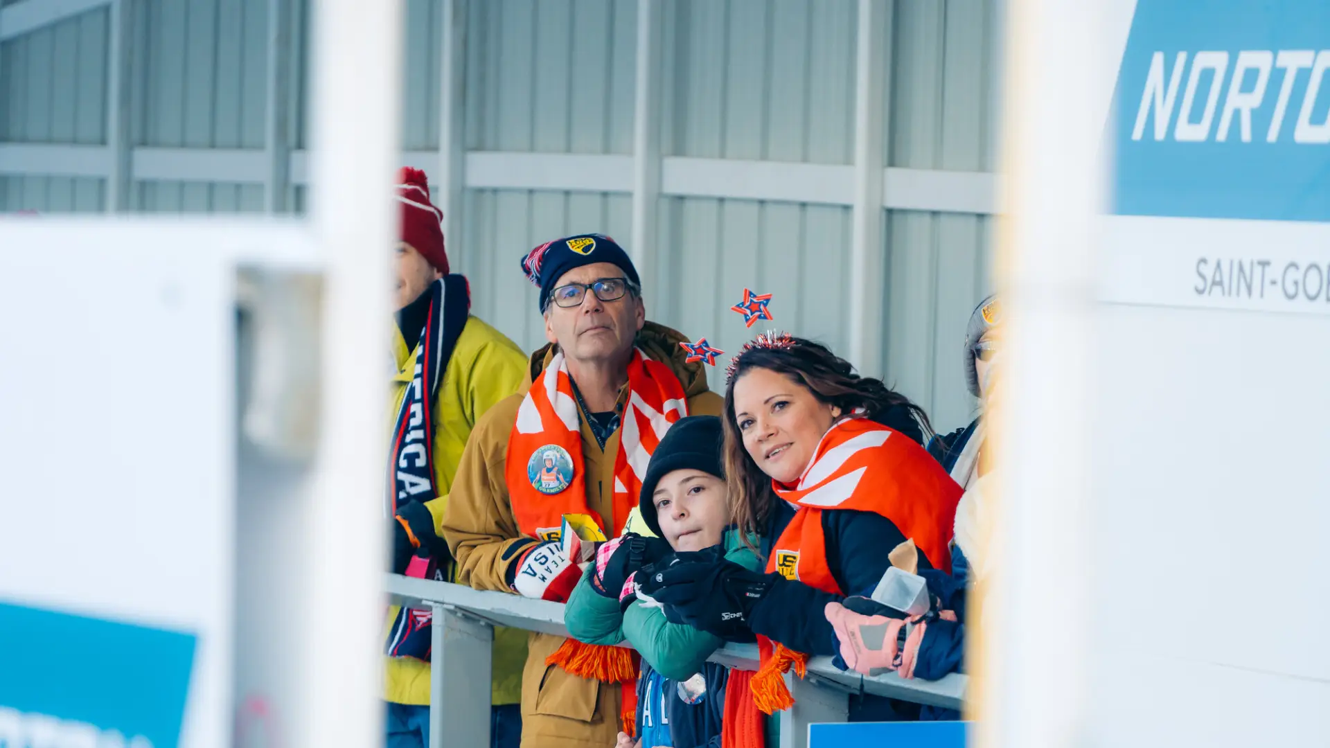 Spectators wearing winter and country-specific attire stand under awning looking onto luge track