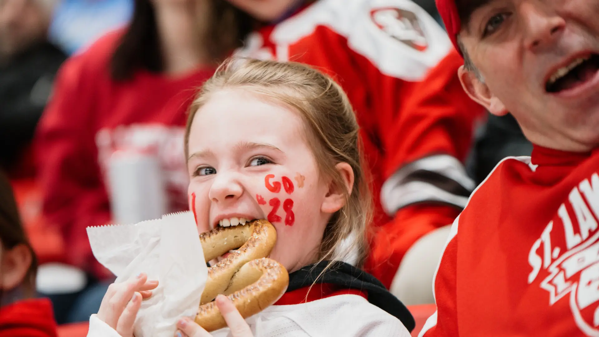 Young girl with blonde hair and "Go 28" painted in red on her cheek looks at the camera as she bites into a large soft pretzel. 