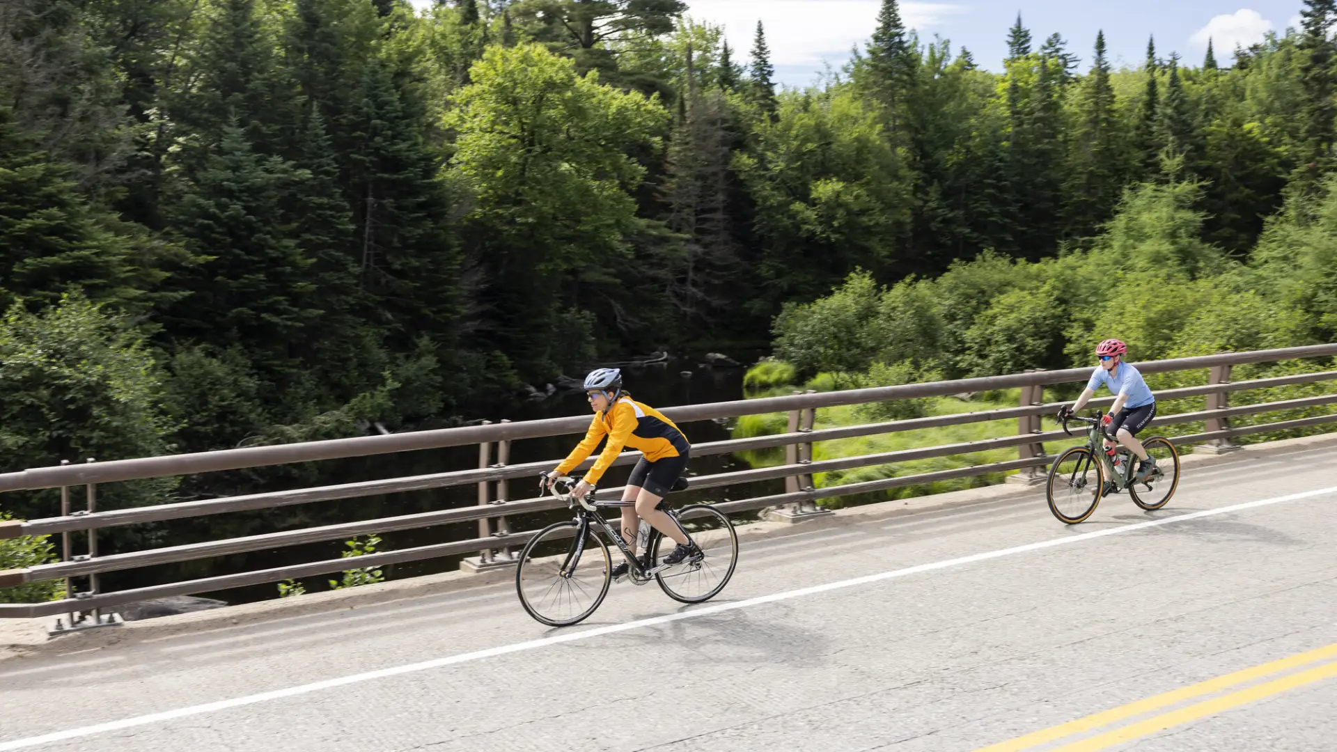 Two cyclists ride on paved bridge with metal railing over river with mix of green trees in background