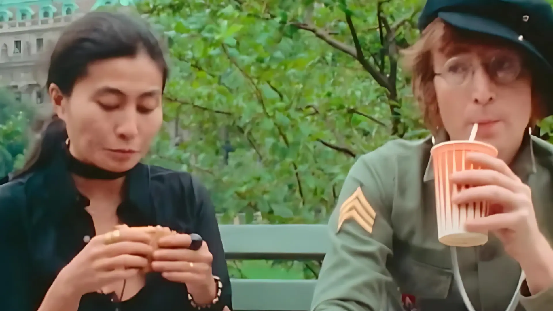 A woman with dark hair eats a sandwich while John Lennon, wearing a newsboy cap and glasses, drinks from a paper cup.  They are sitting on a park bench outdoors.