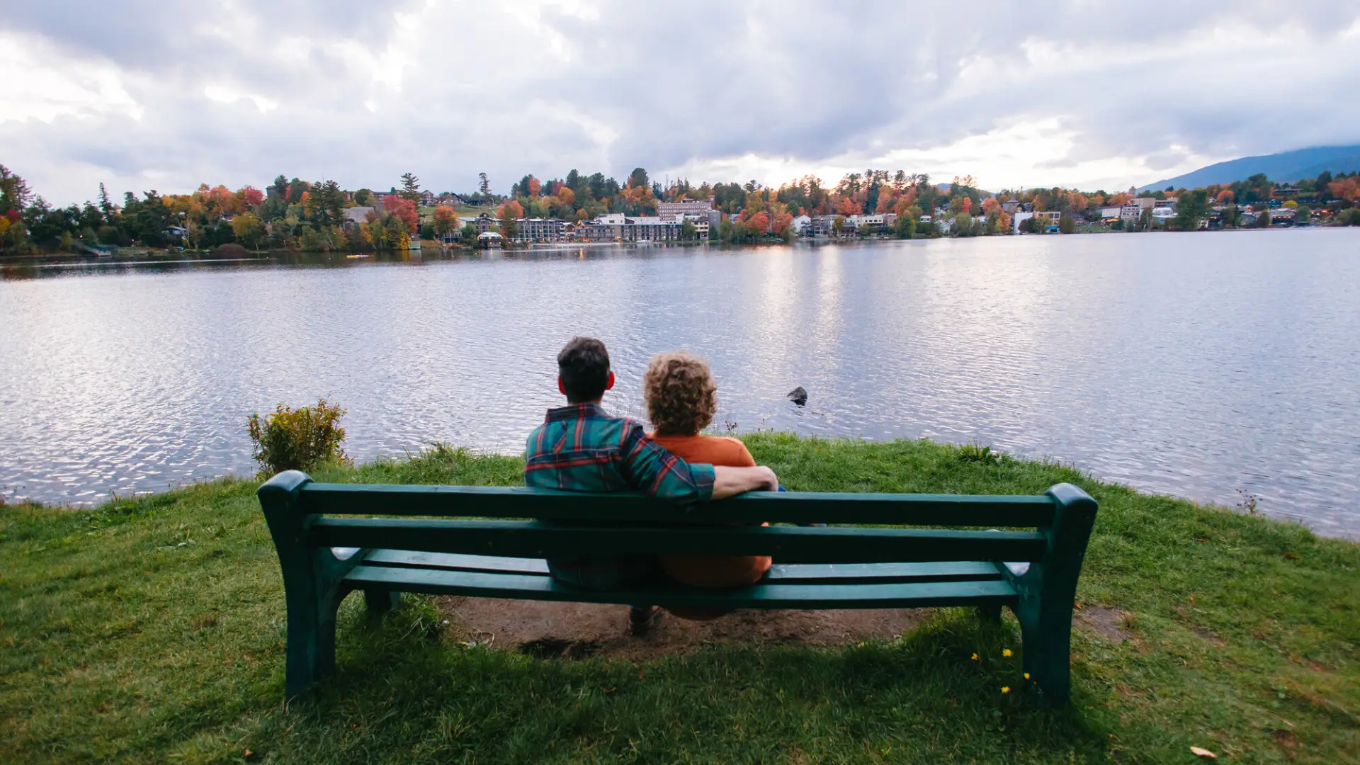 Couple sits on green bench next to lake looking over water at Village of Lake Placid during fall