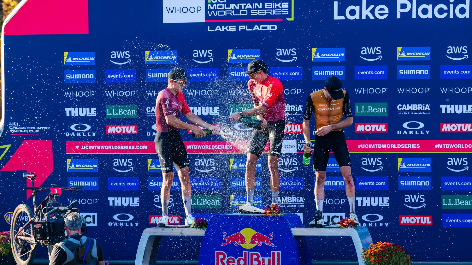 Three mountain bike racers stand atop three stage podium spraying each other with event logos on blue backdrop