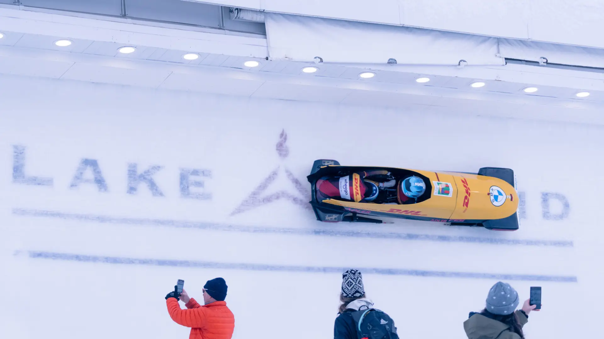 Yellow bobsled descends ice track with Lake Placid logo frozen into ice while people take photos and look on