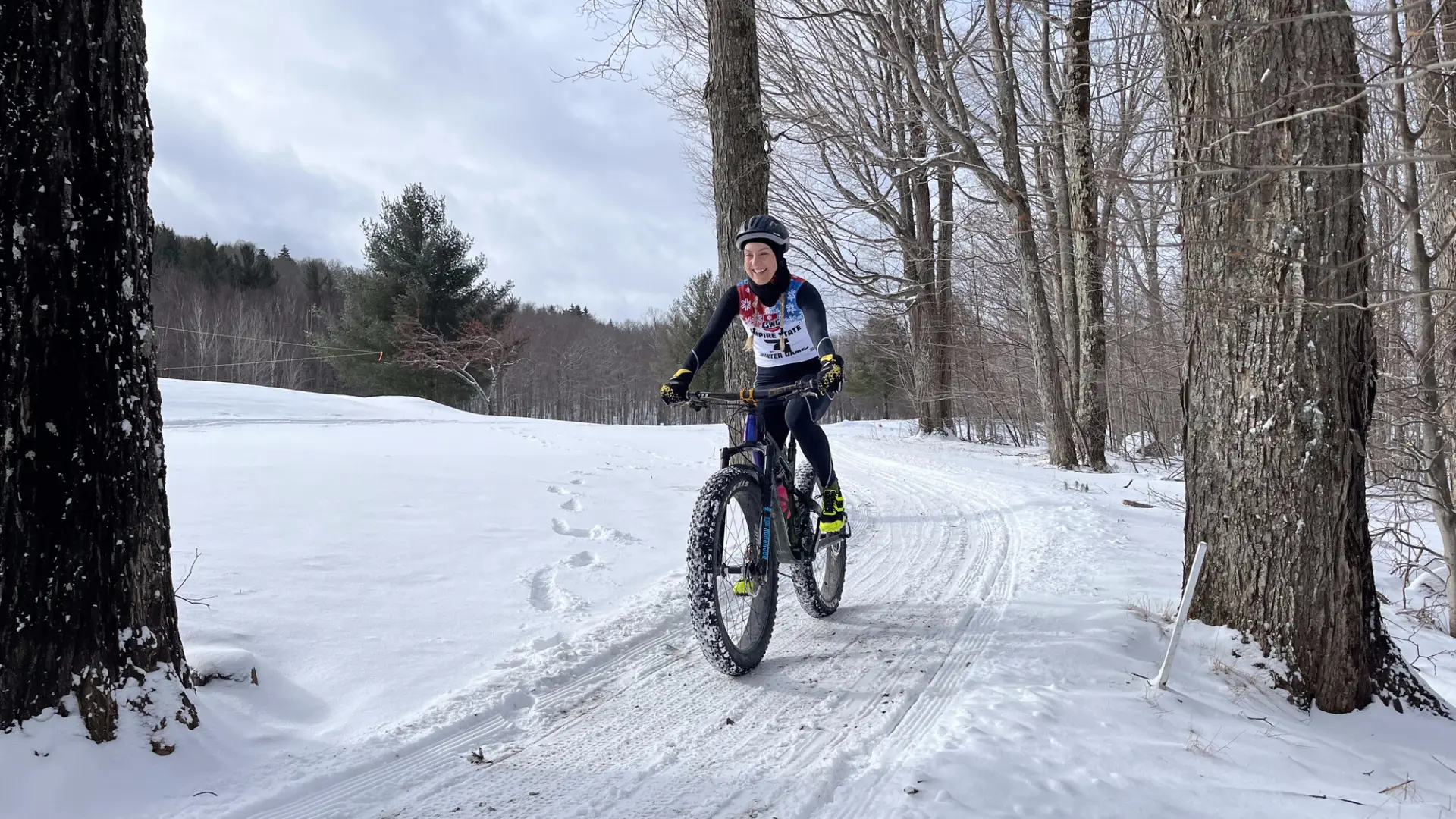 A person smiling while riding a fat-tire mountain bike on a groomed, snowy trail through a wintry forest. 