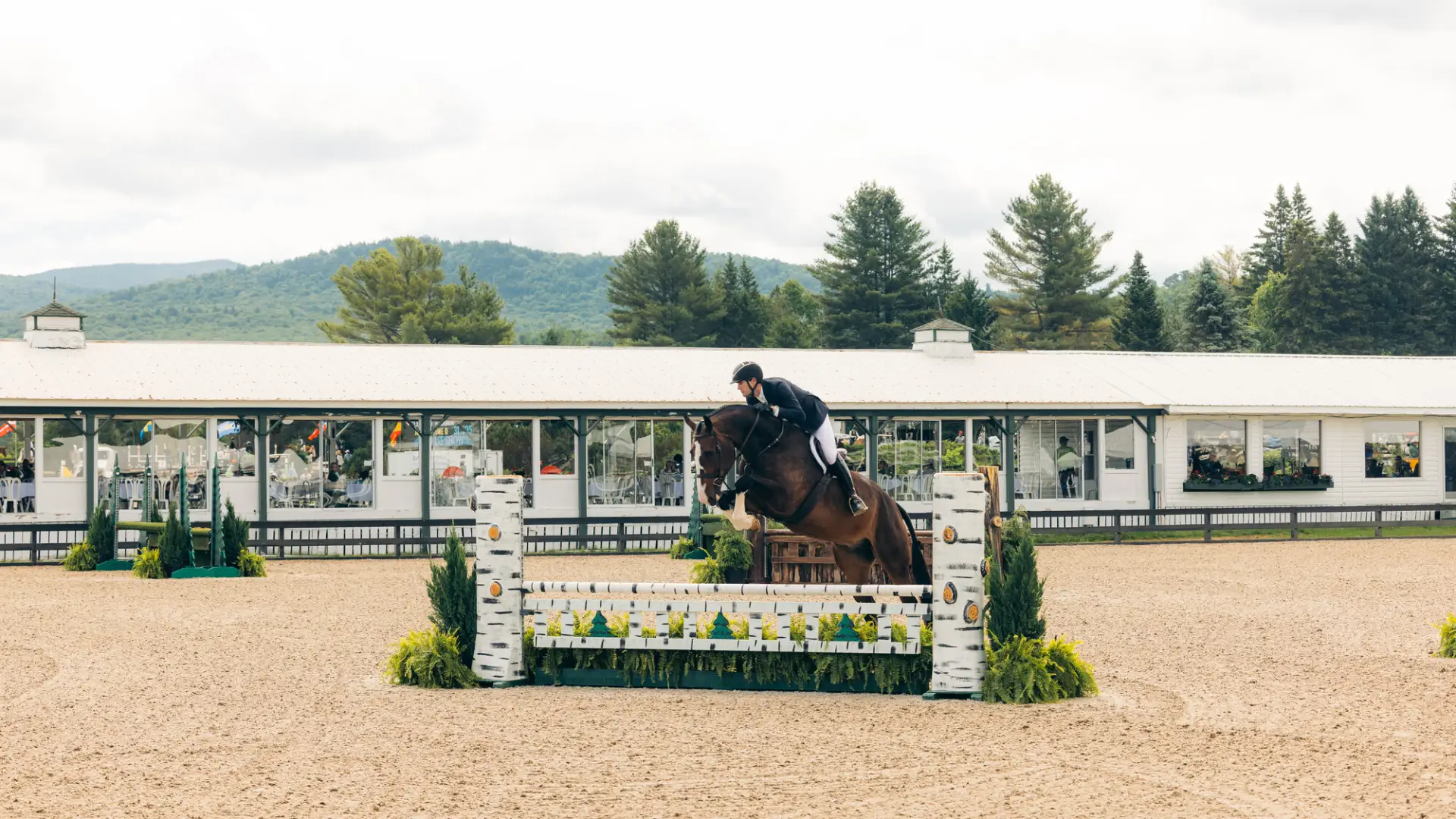 Equestrian rider jumps horse over jump on dirt ring with pavilion building in background