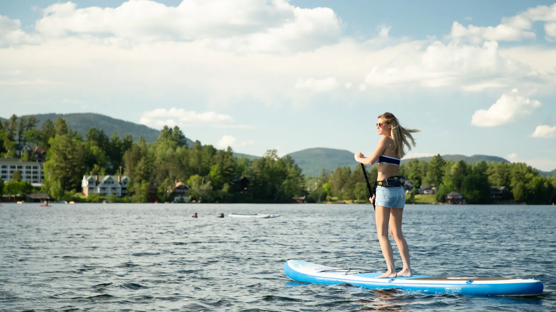 Woman in bathing suit top and shorts wearing sunglasses paddles SUP on lake in summer