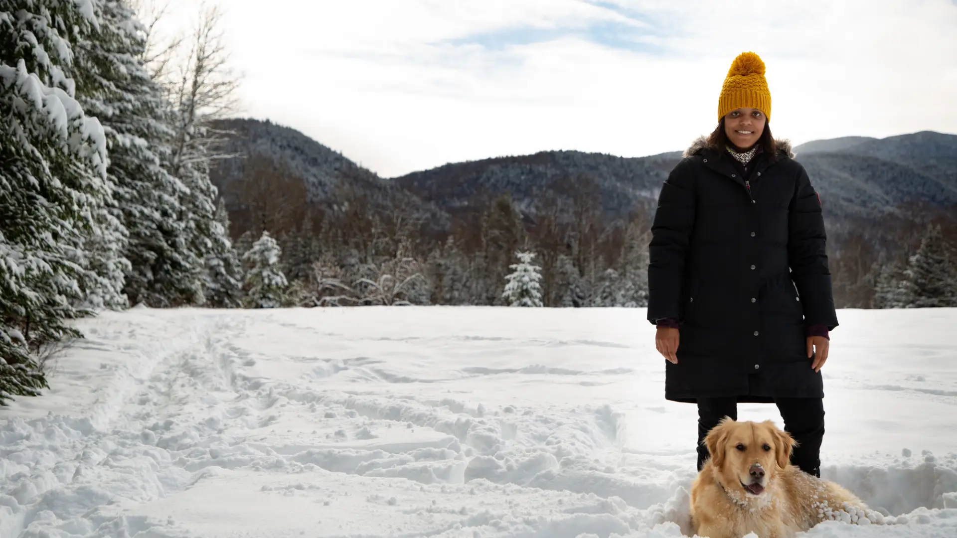 A woman in black winter clothes in a yellow hat poses with her golden retriever in the snow. 
