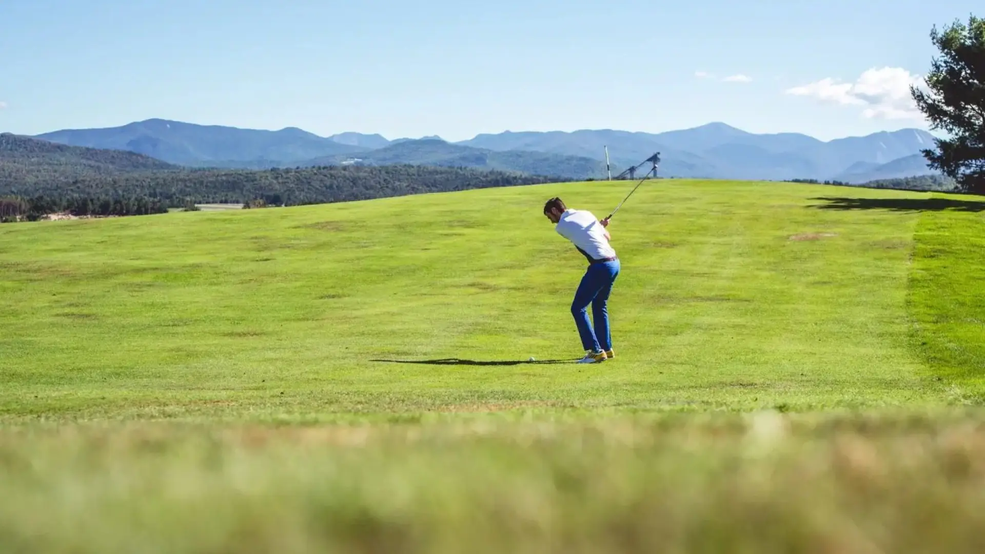 A man on a wide open golf green swings his club and hits the ball toward the ski jumps and mountains in the distance.