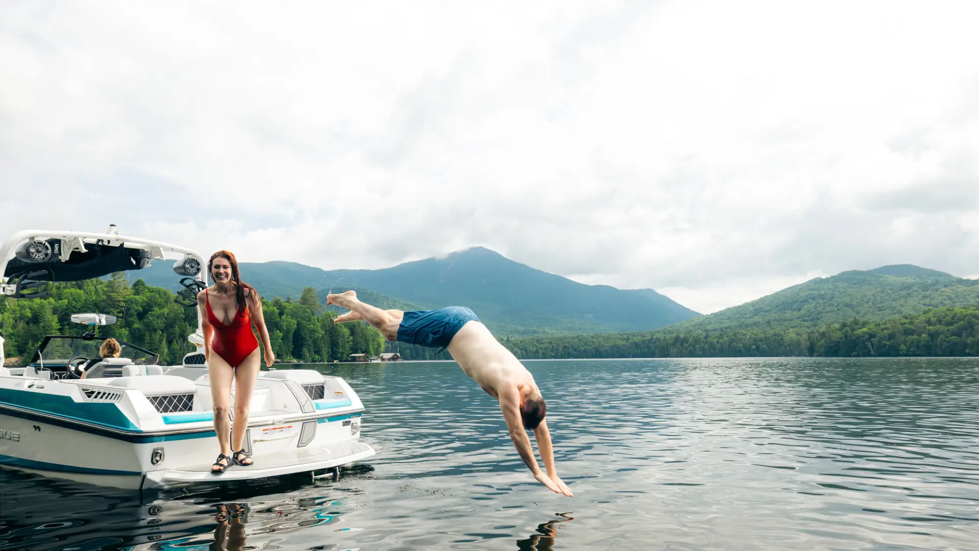 Man dives off motor boat into Lake Placid while woman in red bathing suit looks on laughing with mountains in background