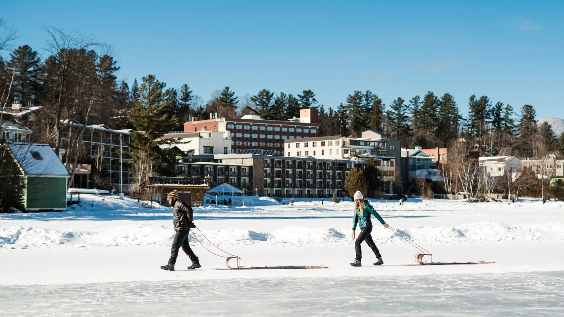 Couple walks across icey lake pulling toboggans with town in the background