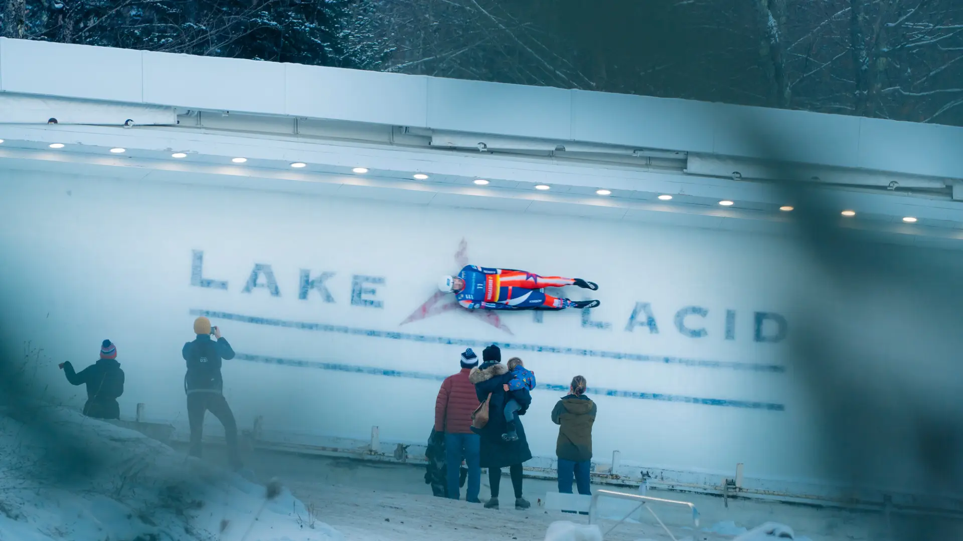 View partially obscured by trees of luge athlete riding down track over where Lake Placid is printed into the ice while several people look on