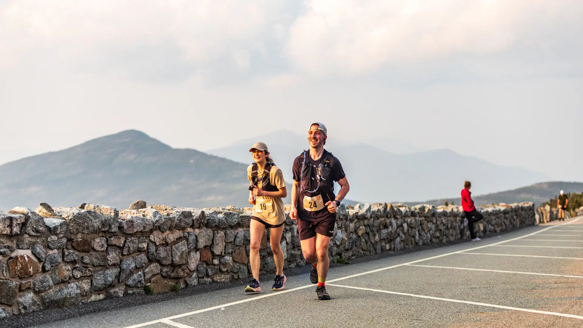 Two smiling runners wearing race bibs and hydration vests jog up a mountain road lined by a stone wall.