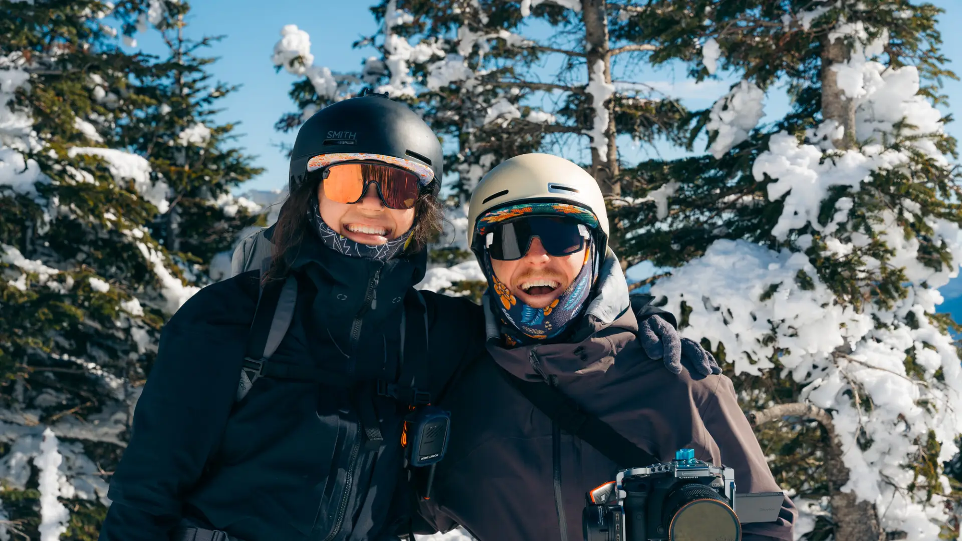 a couple bundled up for adventures in the cold smiles at the camera in front of a wintery scene
