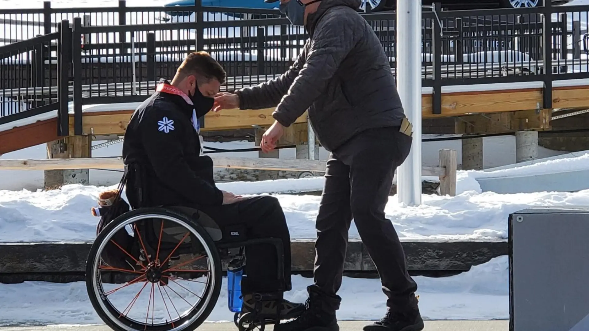 A man stands beside a man in a wheelchair to help him along a sidewalk. Image courtesy of David Christopher.