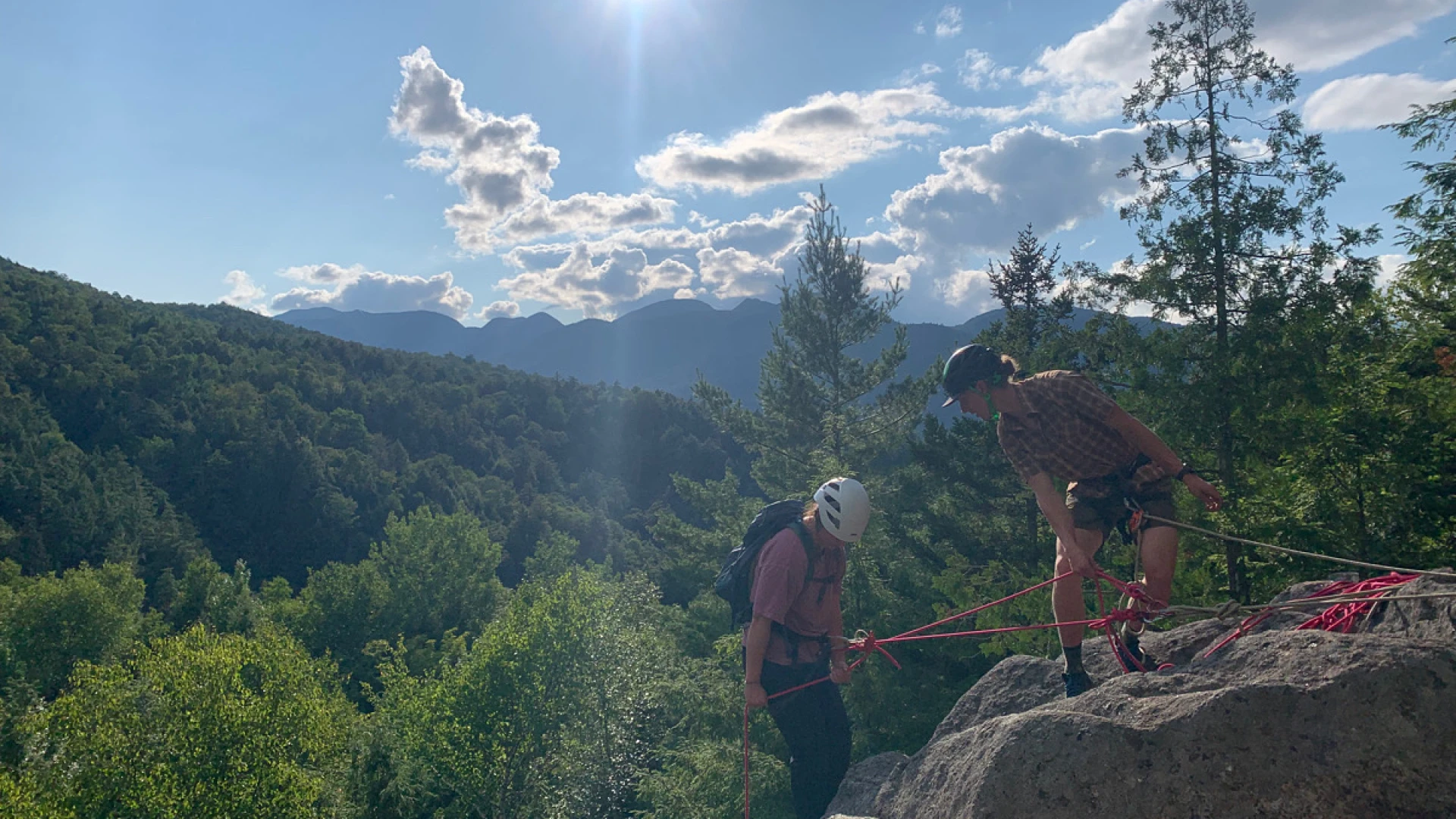 Two rock climbers on a sunny day, one belaying the other on a rocky outcrop overlooking a lush green valley and distant mountains.