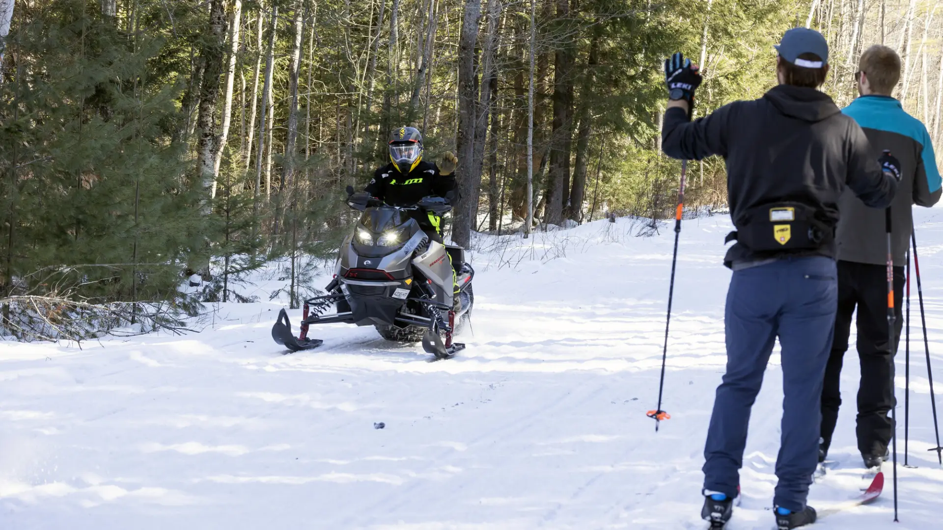 Skiers wave to a snowmobiler on a wide snowy pathway. 