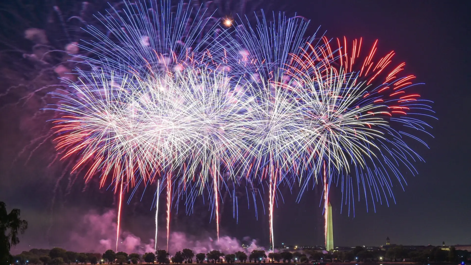 A vibrant fireworks display over a body of water, with the Washington Monument visible in the background.