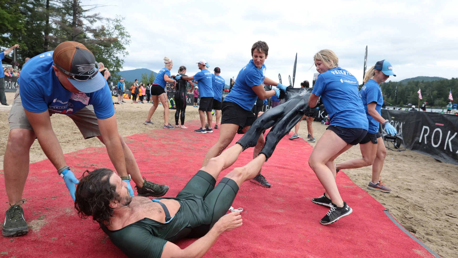 A swimmer lies on a red carpet on the ground while three volunteers in blue Ironman t-shirts remove his wetsuit, one holding his shoulder and two pulling the suit off his legs