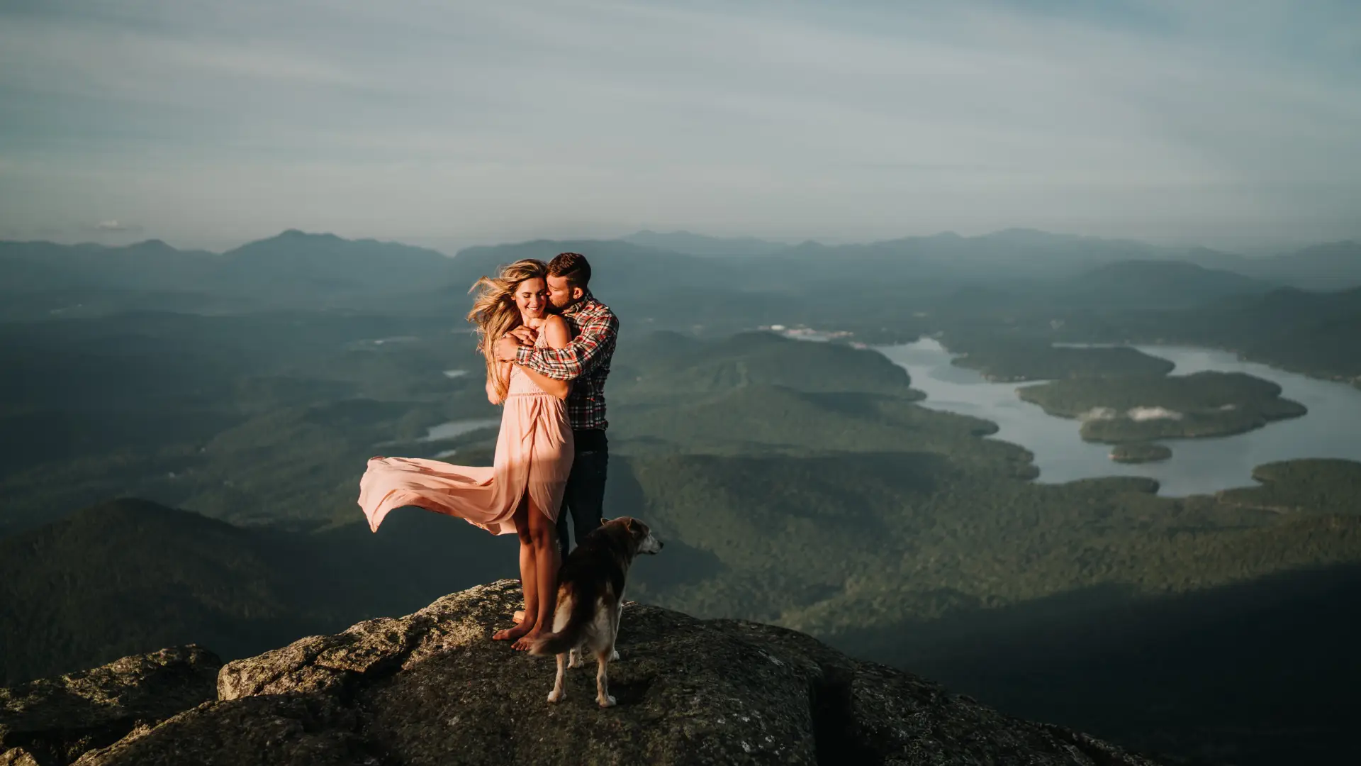 A couple embraces on a rocky ledge while her dress and hair flow in the breeze with Lake Placid in the back drop