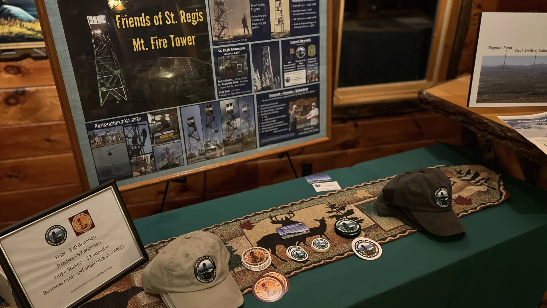 Display table with information about the restoration of the St. Regis Mountain fire tower, including photos and details about its history and construction.  Also shown are hats, patches, and stickers for sale to support the restoration.
