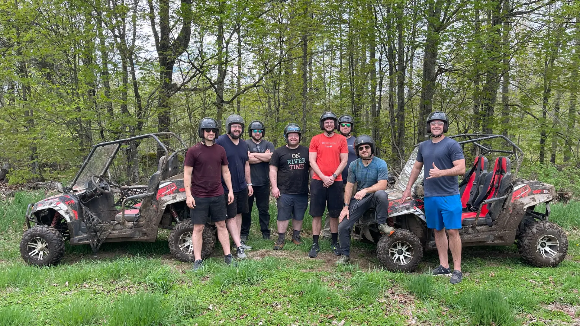 Eight happy young men stand together wearing helmets and smiling between all terrain vehicles in a grassy patch surrounded by trees