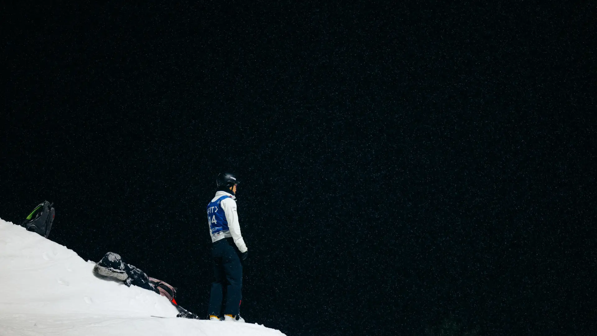 Freestyle aerial skier stands atop a snowy hill on a dark night looking out
