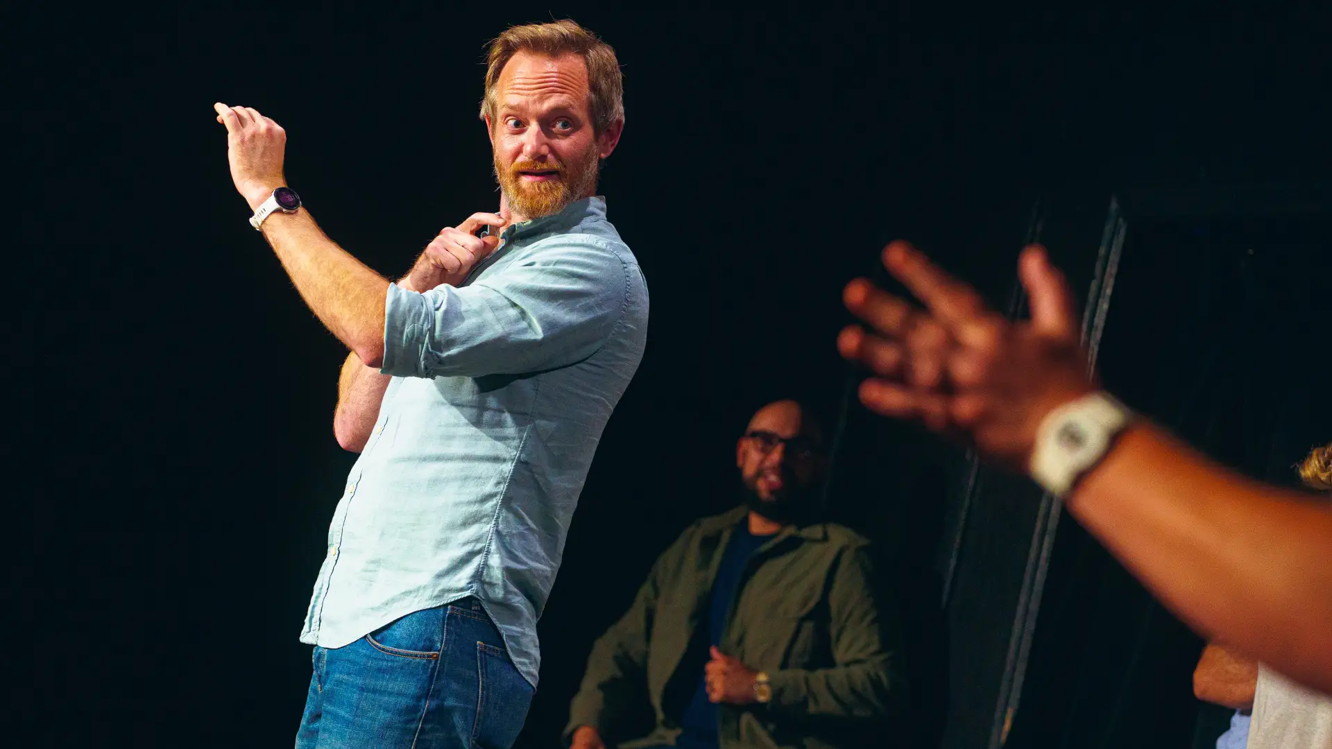 Bearded man in dress shirt and jeans holds up one hand and looks and points in the direction of someone out of image adjacent