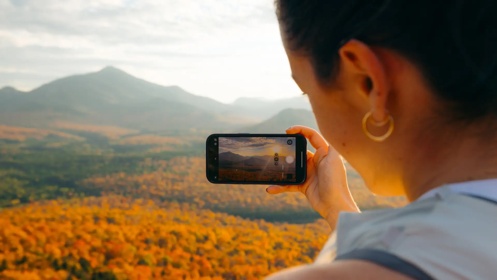 A woman looks at a fall mountain range through a phone camera.